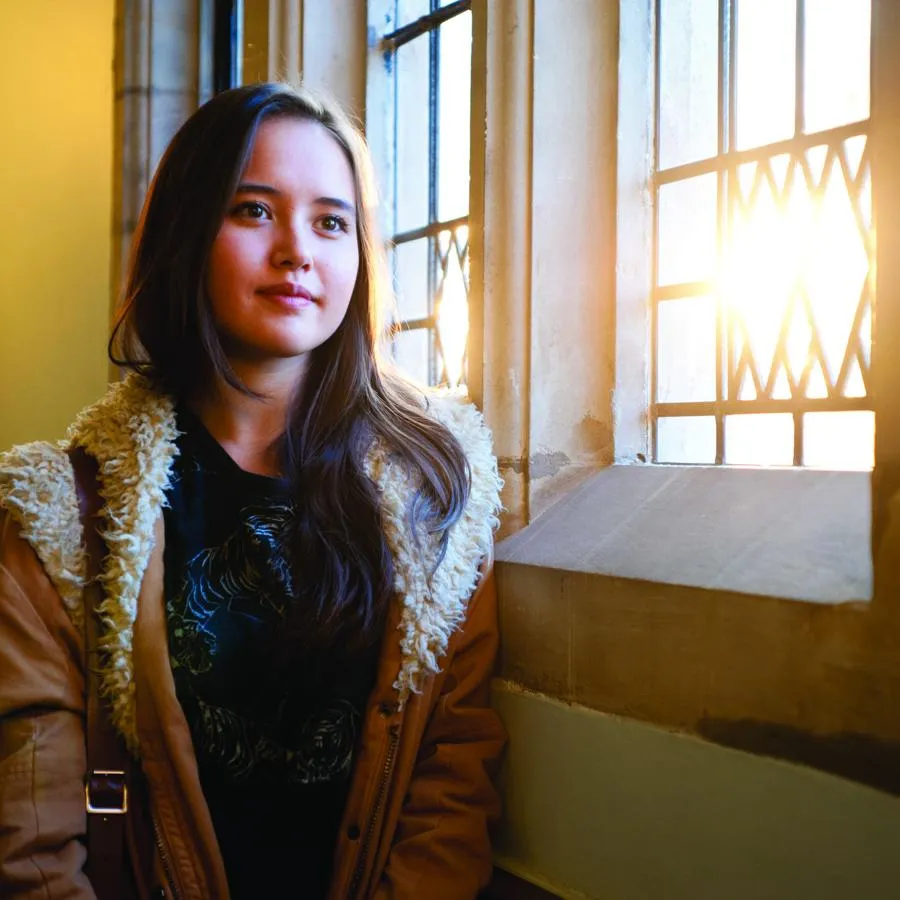 A young woman stands in front of an ornate window in a historic building with the sun streaming in