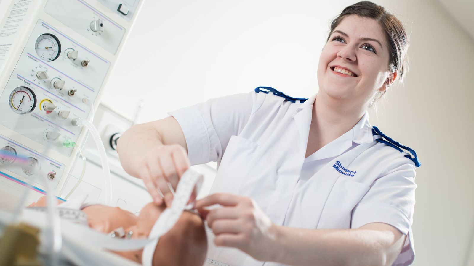 A smiling midwifery student in white NHS uniform practises on a dummy baby
