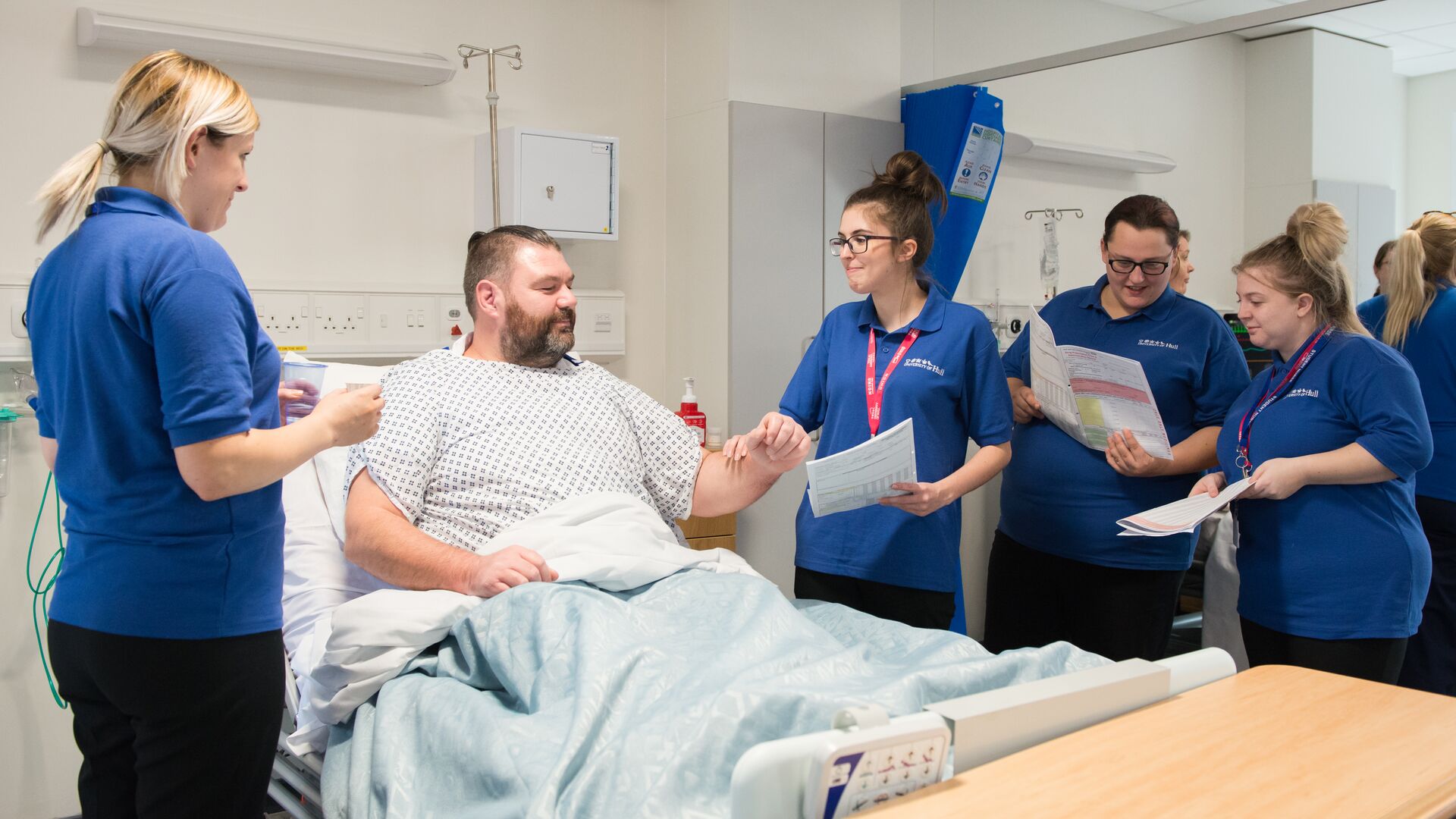 A male patient lays in a hospital bed while nursing students speak to him