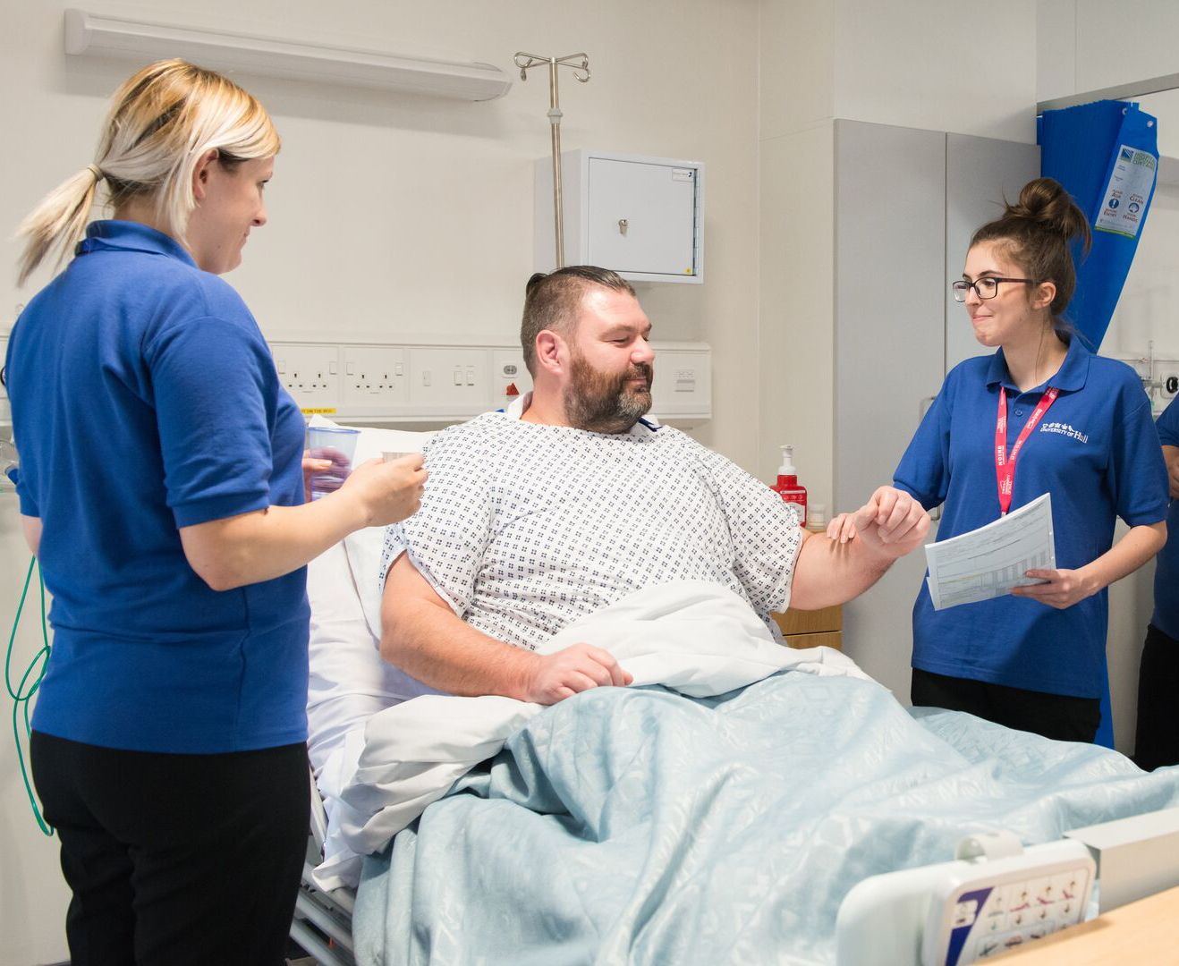 A male patient lays in a hospital bed while nursing students speak to him