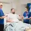 A male patient lays in a hospital bed while nursing students speak to him