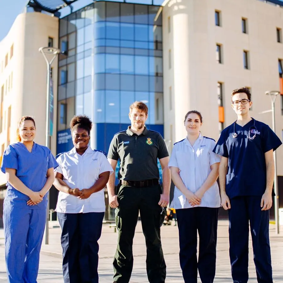 A group of five health students from various fields standing in their respective uniforms in front of health training building on campus