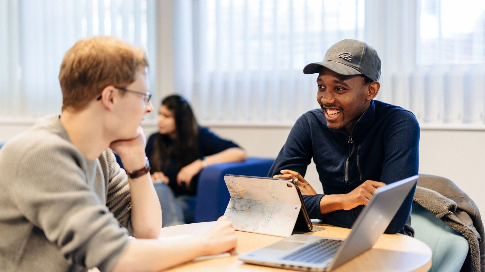 Two students sitting facing each other at a table with a laptop and tablet on the desk
