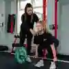 Two students in a gym, one student is lifting a barbell whilst the other is spotting her.