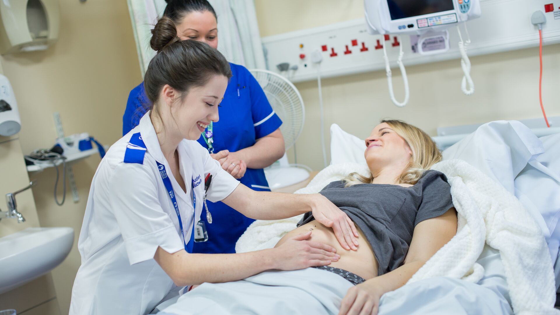 A smiling midwifery student in white NHS uniform examines a pregnant woman while a supervisor watches on