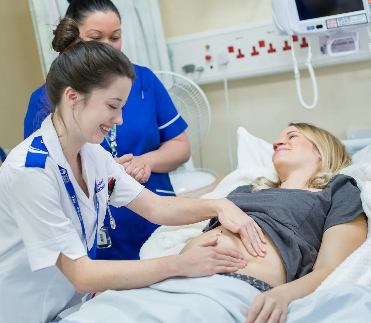 A smiling midwifery student in white NHS uniform examines a pregnant woman while a supervisor watches on