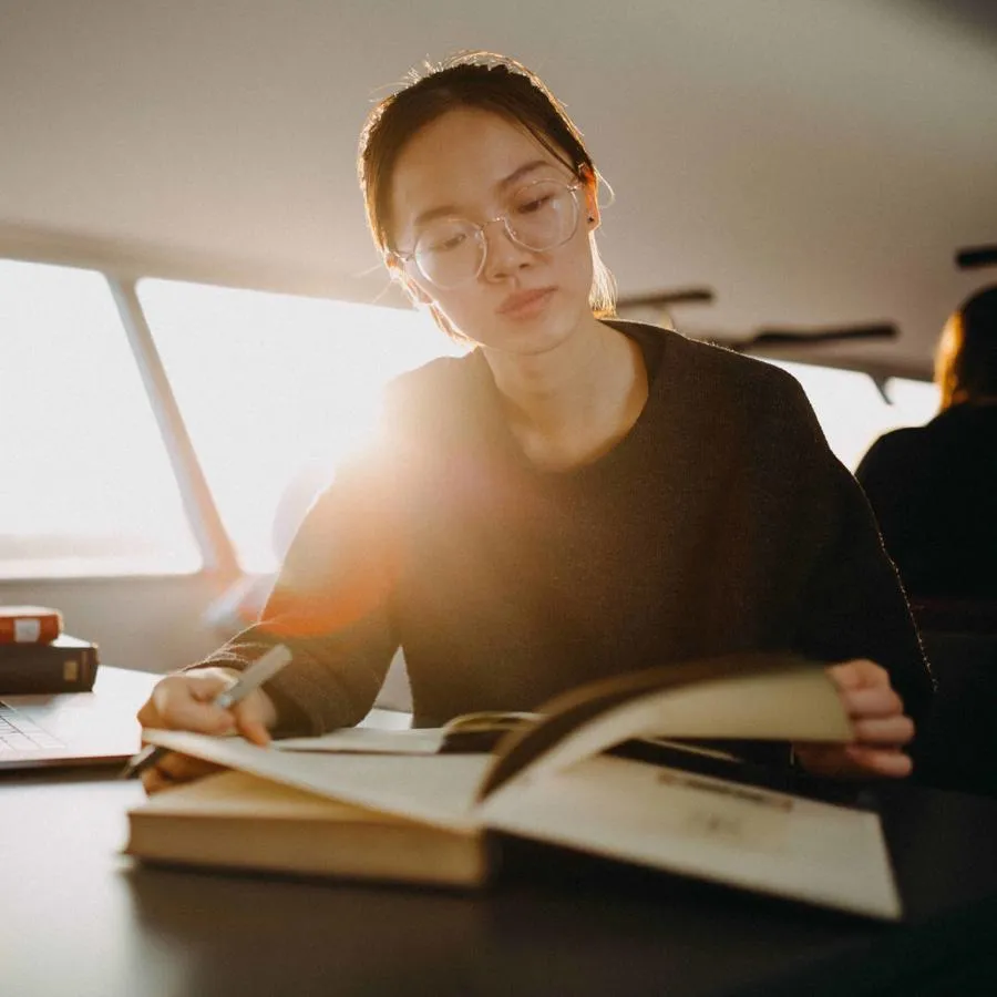 A young woman with glasses reads a book at a desk in a dimly lit room with the sun streaming in behind