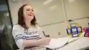 A female maths student in a white jumping laughing while writing in a notebook 