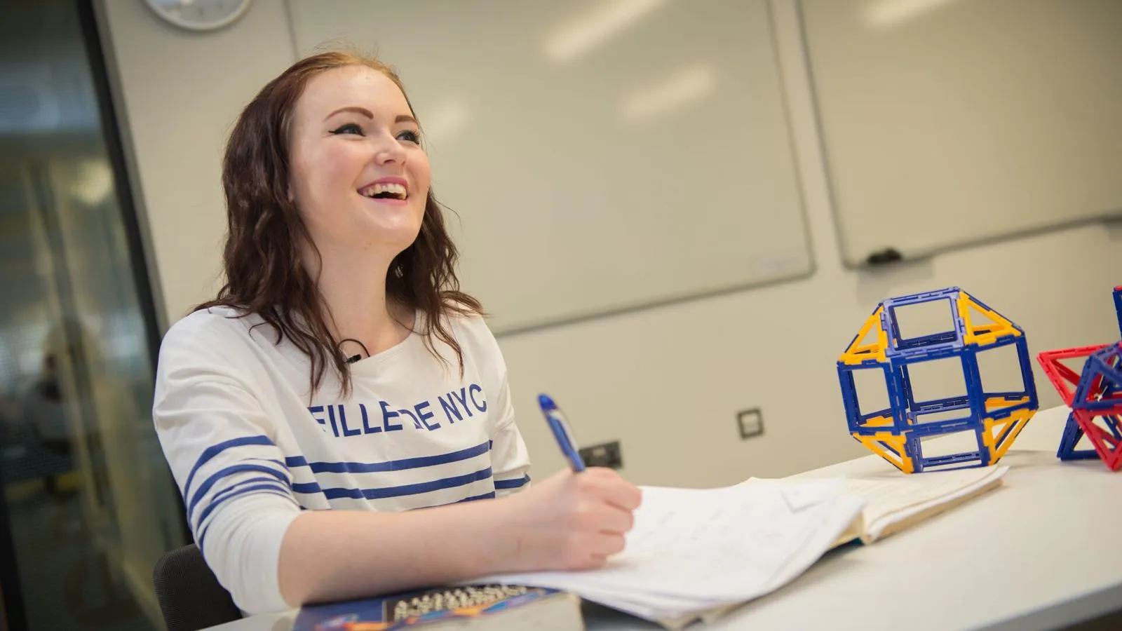 A female maths student in a white jumping laughing while writing in a notebook 