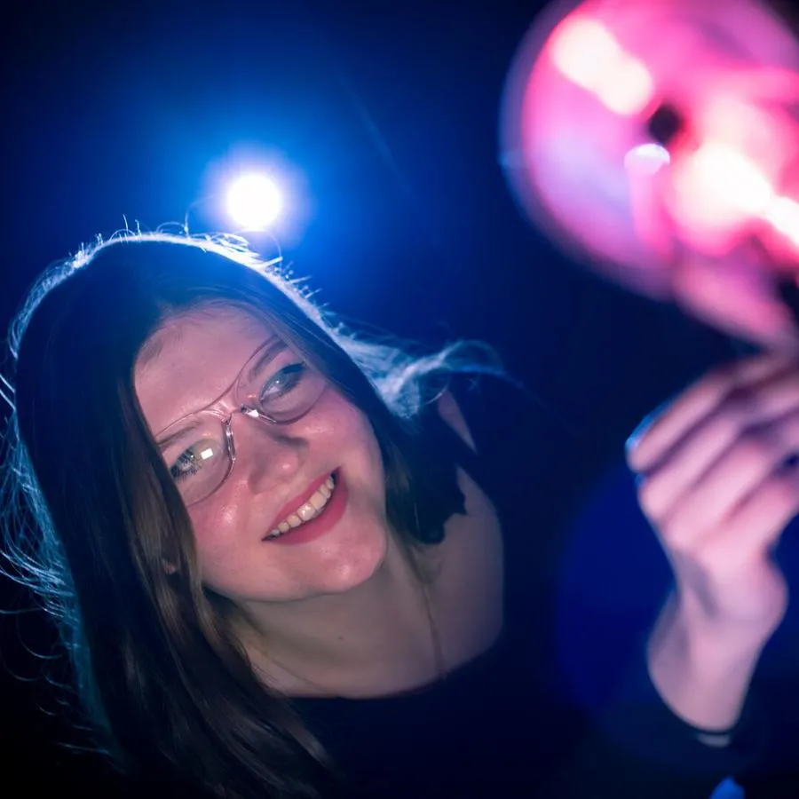 A young woman with dark hair and protective glasses works with brightly coloured equipment in a dark room