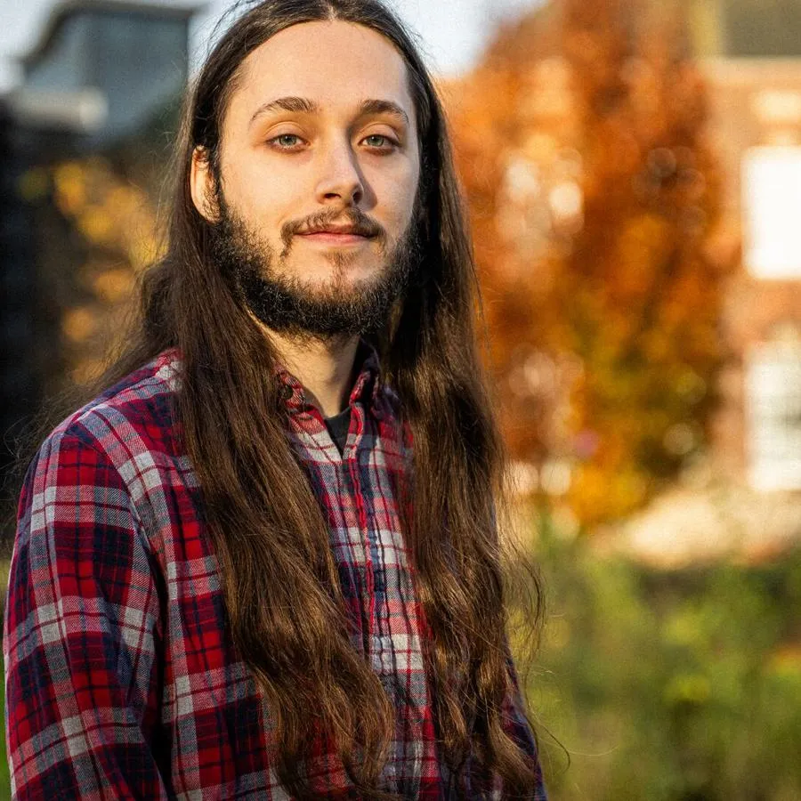 A young man in a red checked shirt standing in front of a red-brick building with autumnal trees in front