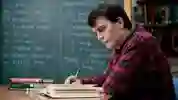 Student in a red checked shirt reading a textbook in front of a chalkboard