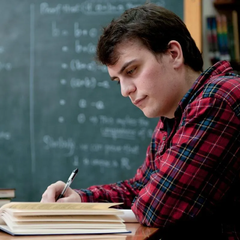 Student in a red checked shirt reading a textbook in front of a chalkboard