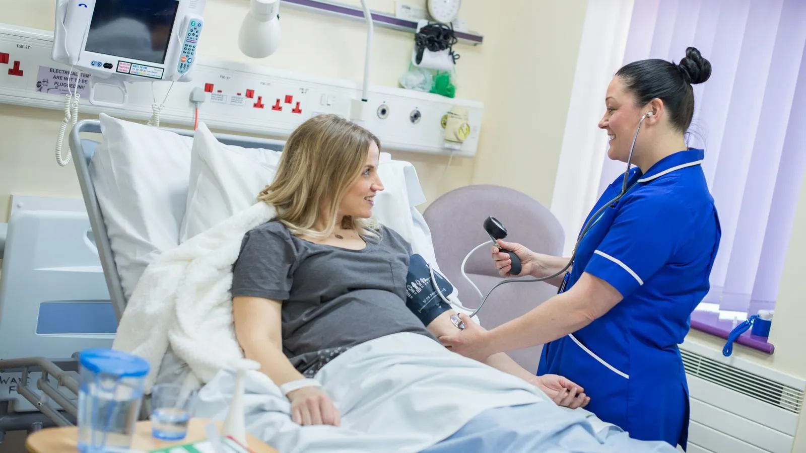 A friendly midwife in blue NHS uniform examines a woman in a hospital bed