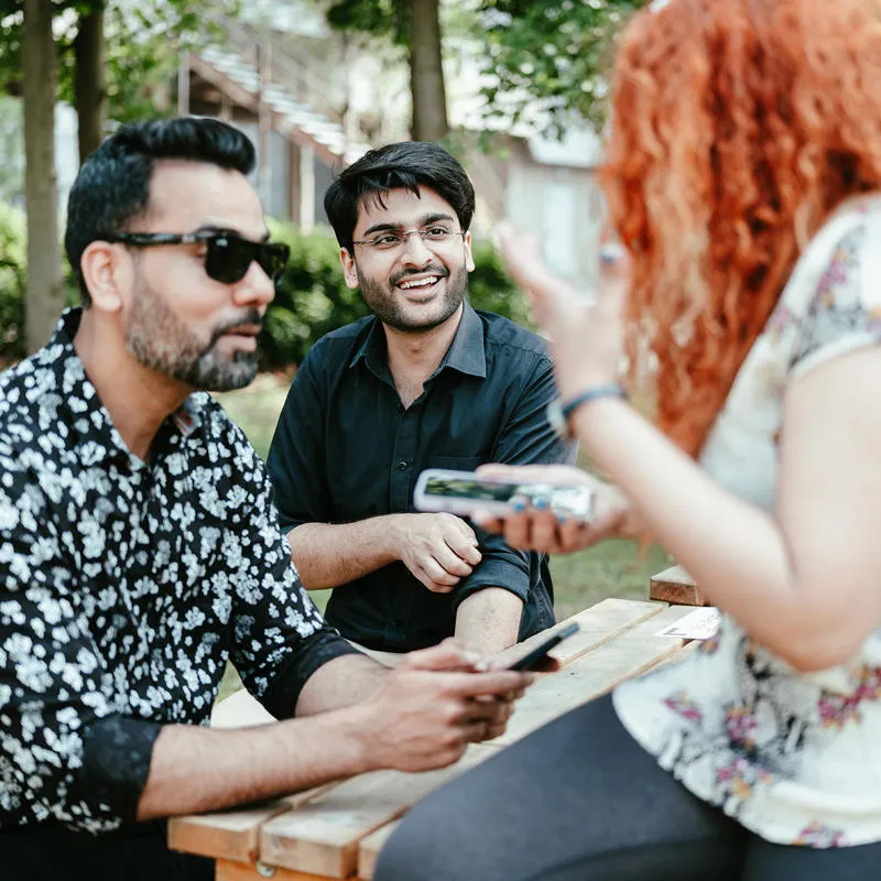 Three students sit at a picnic bench smiling and laughing outside the students' union.