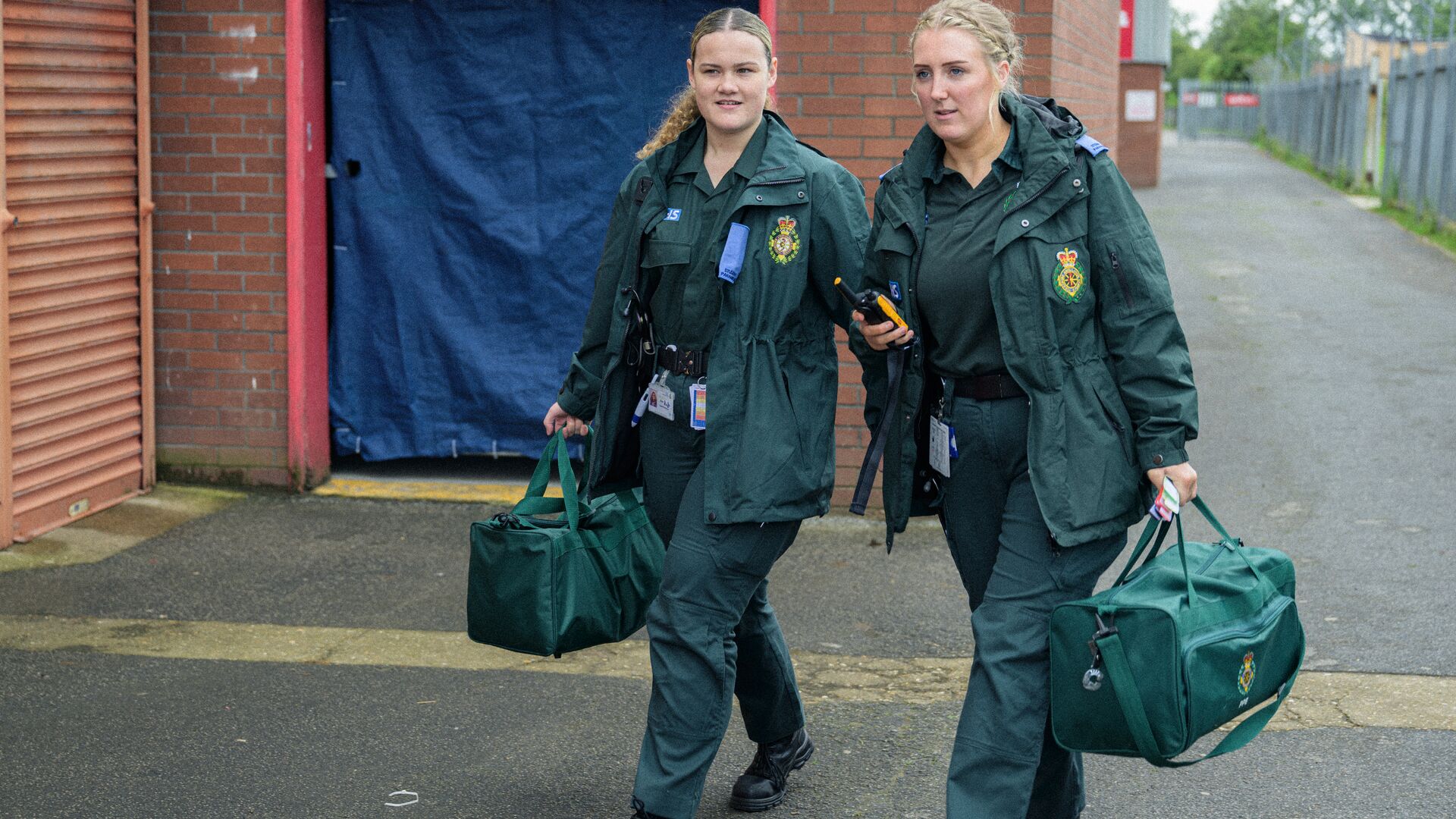Two female student paramedics in green NHS uniform carrying equipment