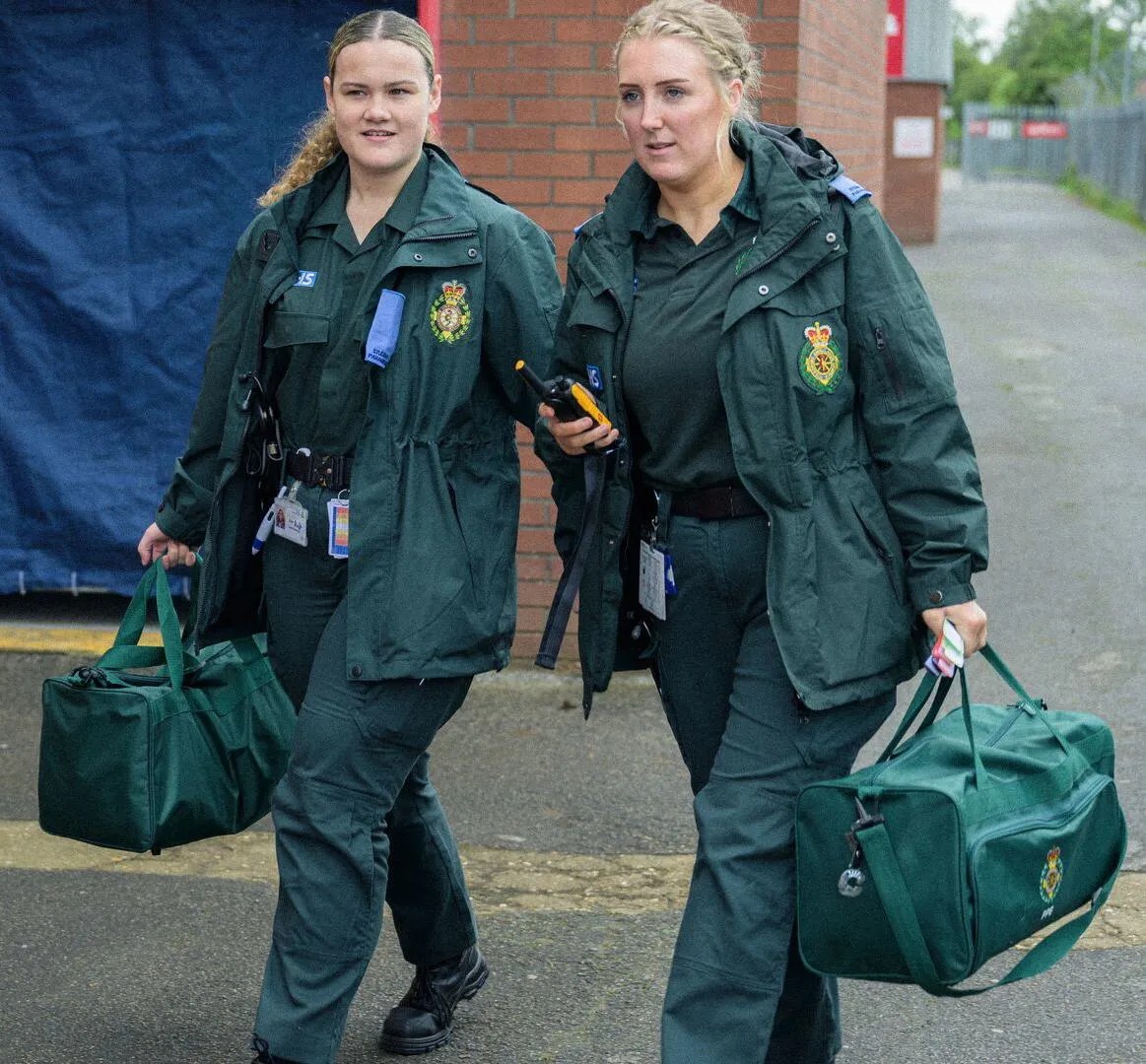 Two female student paramedics in green NHS uniform carrying equipment