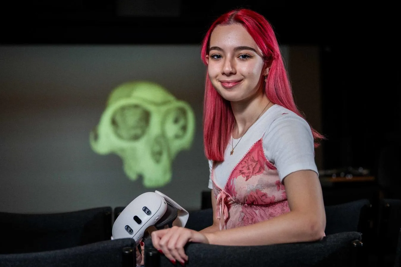 Red haired female student holding VR headset standing in front of image of green skull