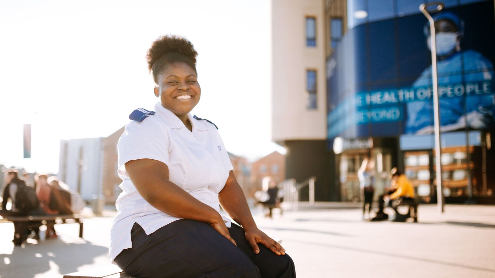 An Adult Nursing student in a white uniform sitting in front of the university's health building on campus