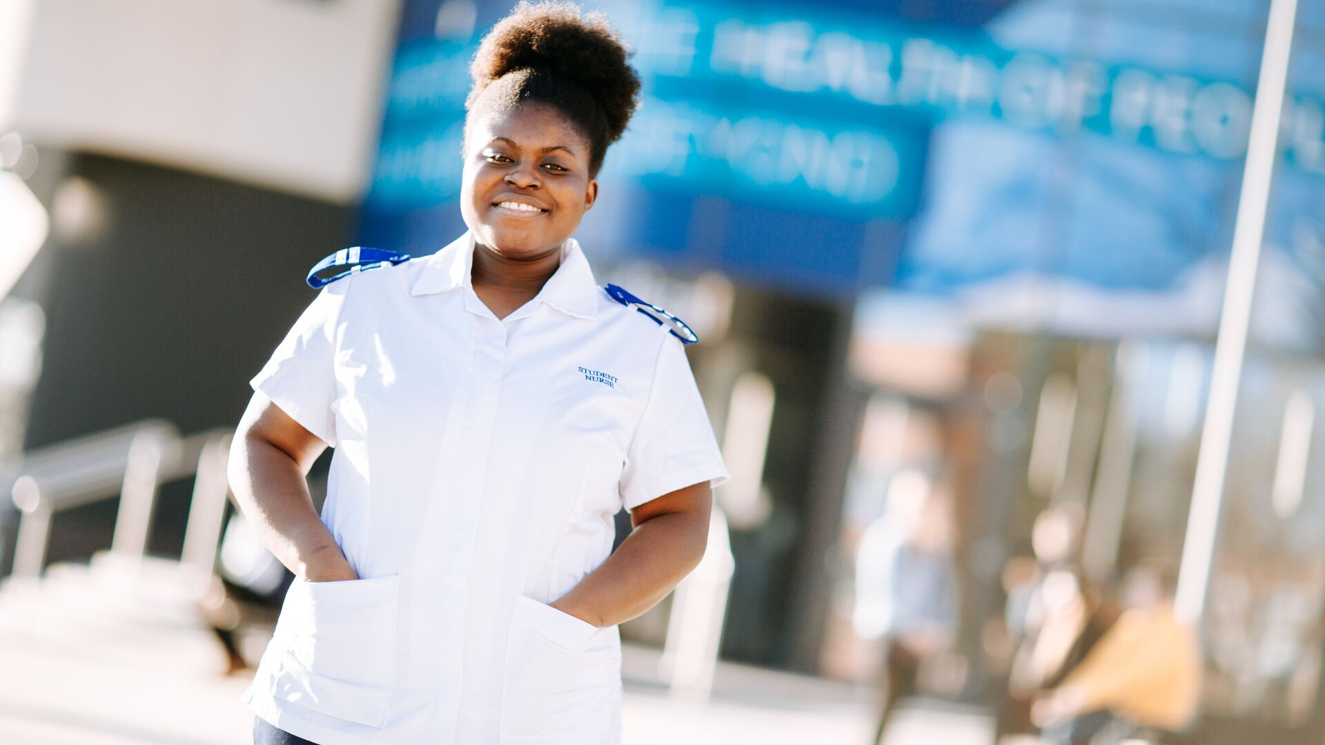 An Adult Nursing student in a white uniform standing in front of the university's health building on campus