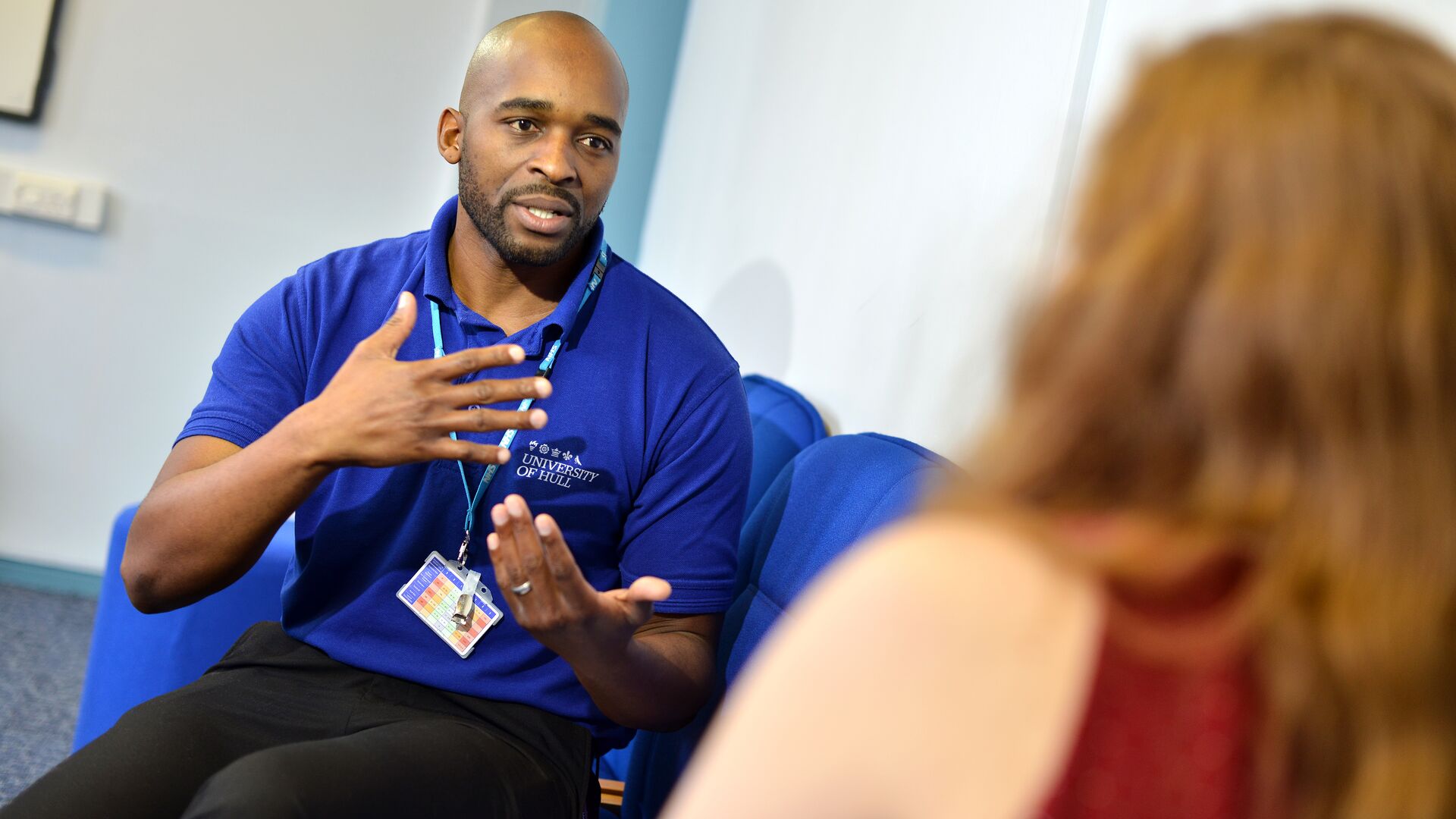 A mental health nursing student in a blue polo shirt speaking to a patient on a sofa