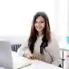 Student in a white room smiles at camera with laptop.