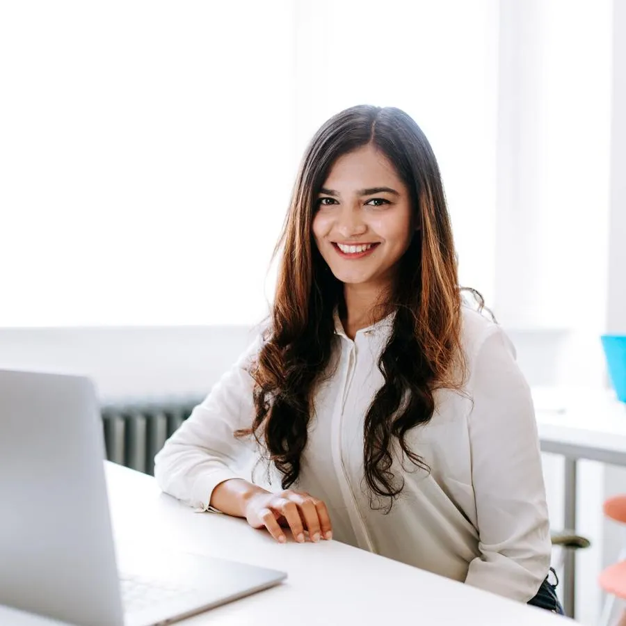 A smiling young woman with long hair sits at a desk with a laptop in a white room