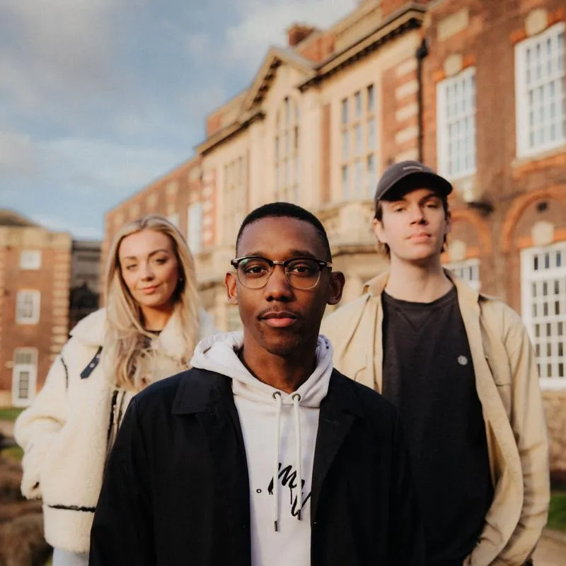 Three of Hull’s students stand confidently together on a lawn in front of beautiful red brick buildings