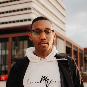 A male student in white hoodie and black jacket stands confidently in front of the Brynmor Jones Library