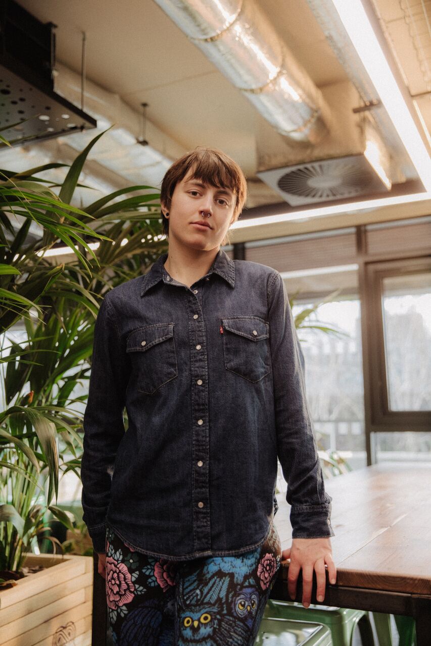 A female student in patterned leggings perches confidently by a table in front of foliage in student hangout The Nest