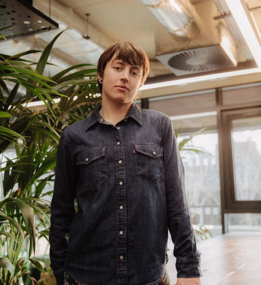 A female student in patterned leggings perches confidently by a table in front of foliage in student hangout The Nest