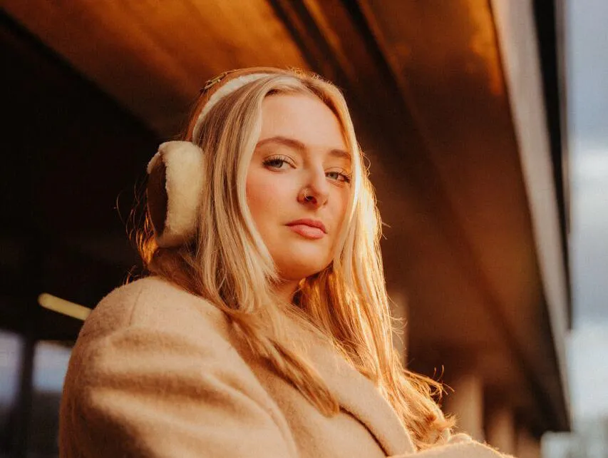A female student in a beige coat with matching earmuffs stands confidently in dramatic sunlight outside the students' union