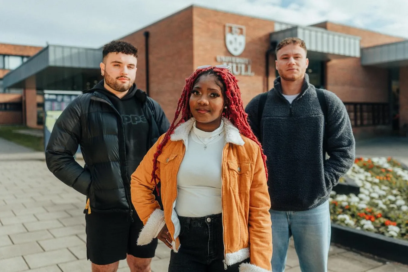 Three of Hull’s students stand confidently together in front of flower beds at the front of campus