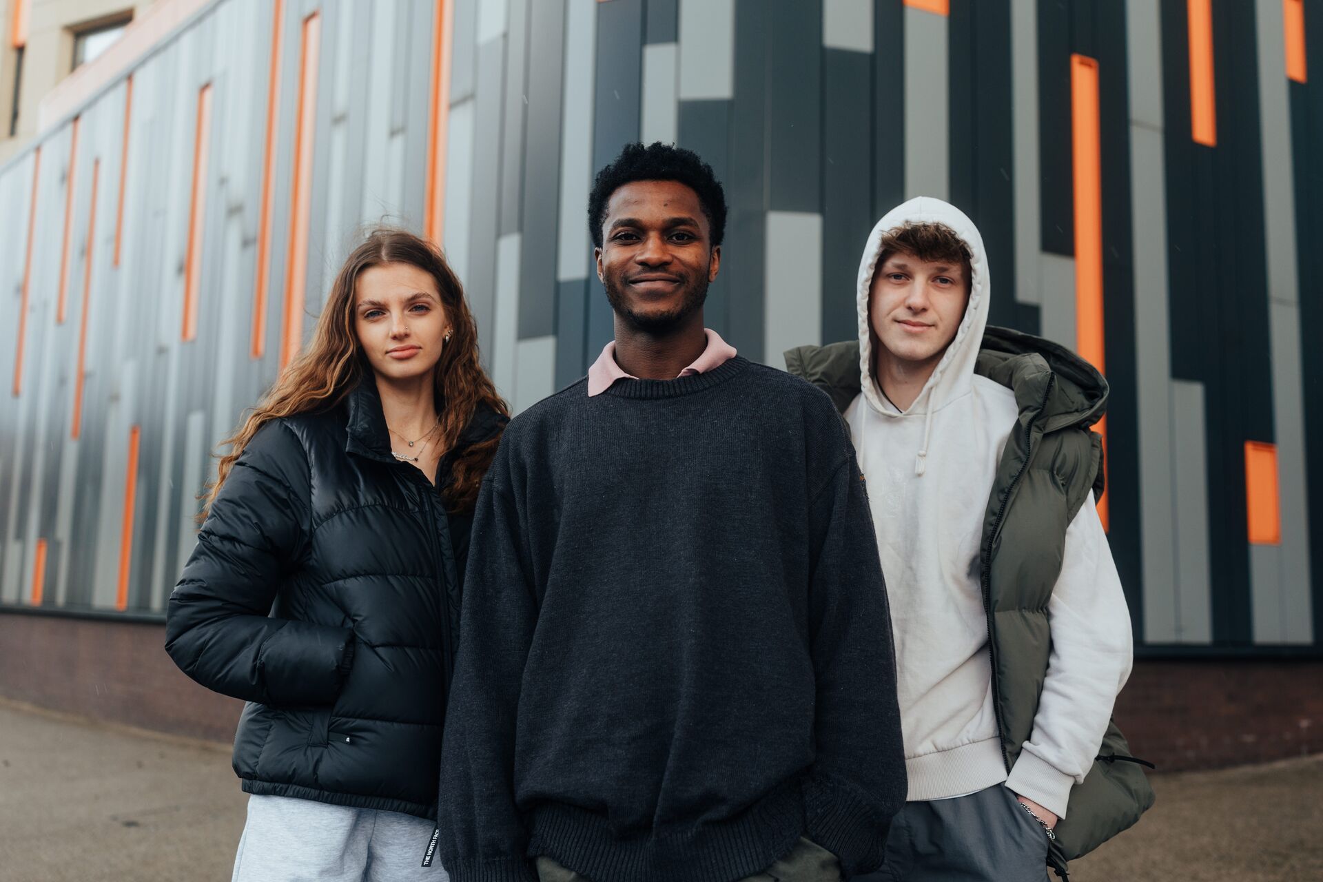 Three of Hull’s students stand confidently together in front of a the exterior black, grey and orange panelled wall of the Medical Building