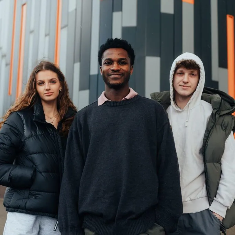 Three of Hull’s students stand confidently together in front of a the exterior black, grey and orange panelled wall of the Medical Building