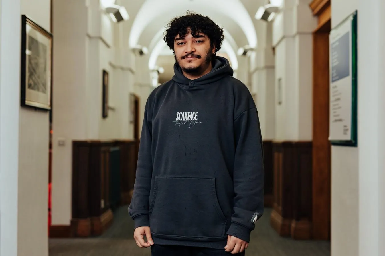 A male student in black hoodie stands confidently in a brightly lit corridor in the business school
