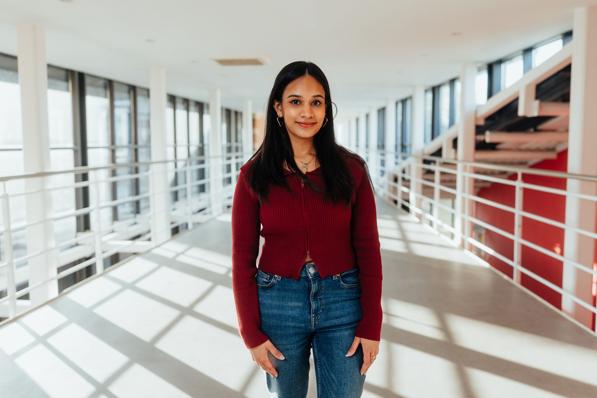 A female student in red sweater and jeans stands confidently on an elevated walkway with red walls in the Business School