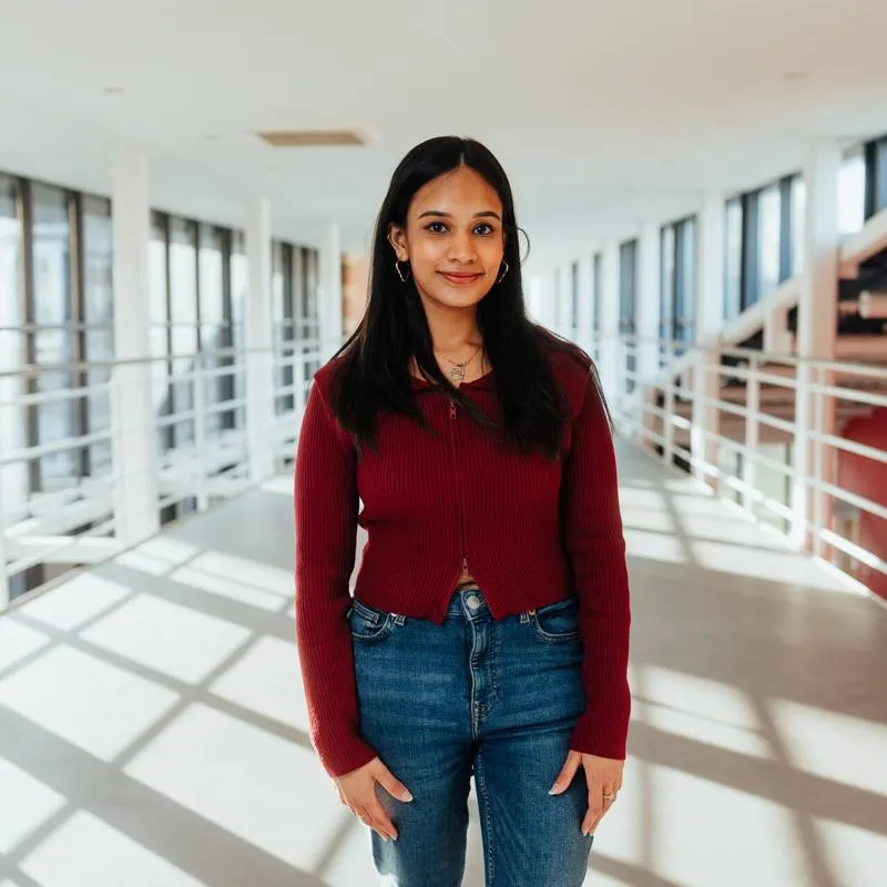 A female student in red sweater and jeans stands confidently on an elevated walkway with red walls in the Business School