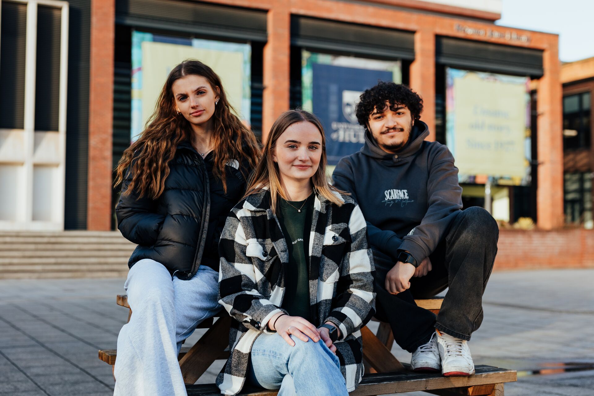 Three of Hull’s students sit confidently together on a picnic table in the Library Plaza