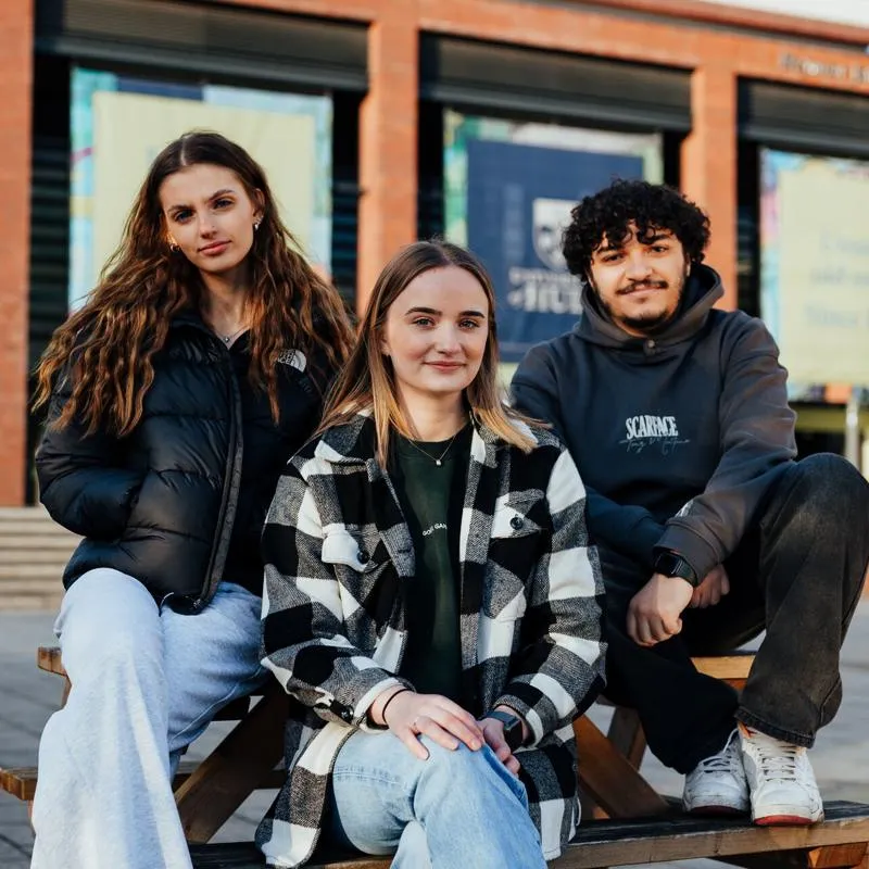 Three of Hull’s students sit confidently together on a picnic table in the Library Plaza