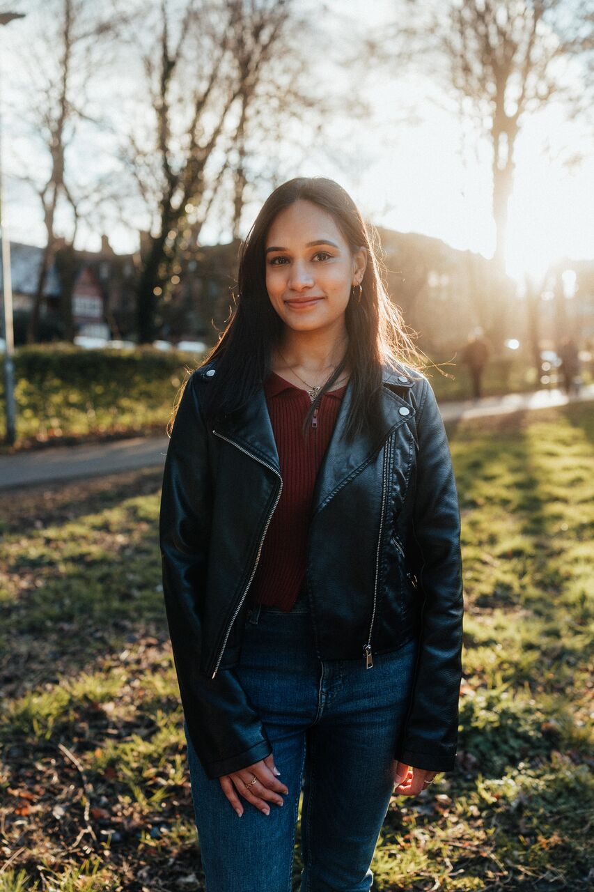 A femal student in leather jacket and red jumper stands confidently under a setting sun in a wooded area of campus