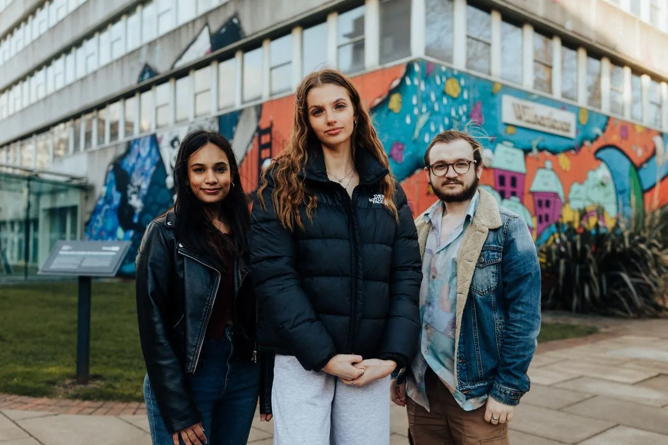 Three of Hull’s students stand confidently together in front of a bright mural on the concrete Wilberforce building