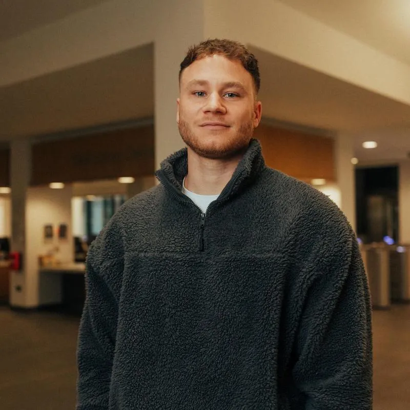 A male student in jeans and fleece stands confidently in front of the Library reception desk