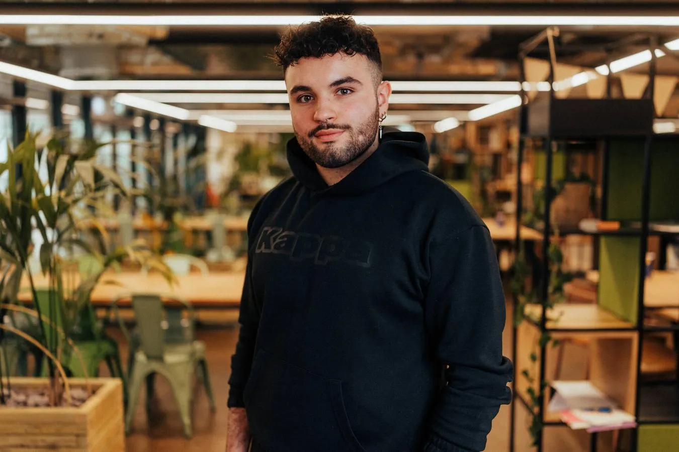 A male student in black hoodie and shorts stands confidently in front of modern lighting and foliage in a student hangout