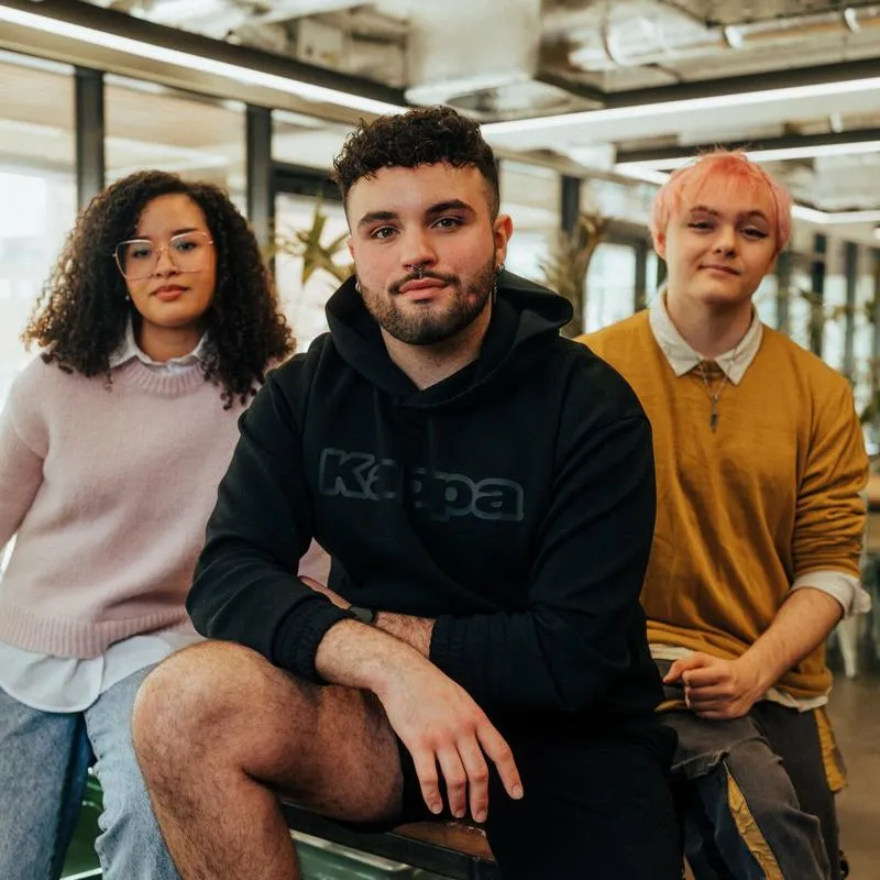 Three of Hull’s students sit confidently together in front of modern lighting and foliage in the student hangout