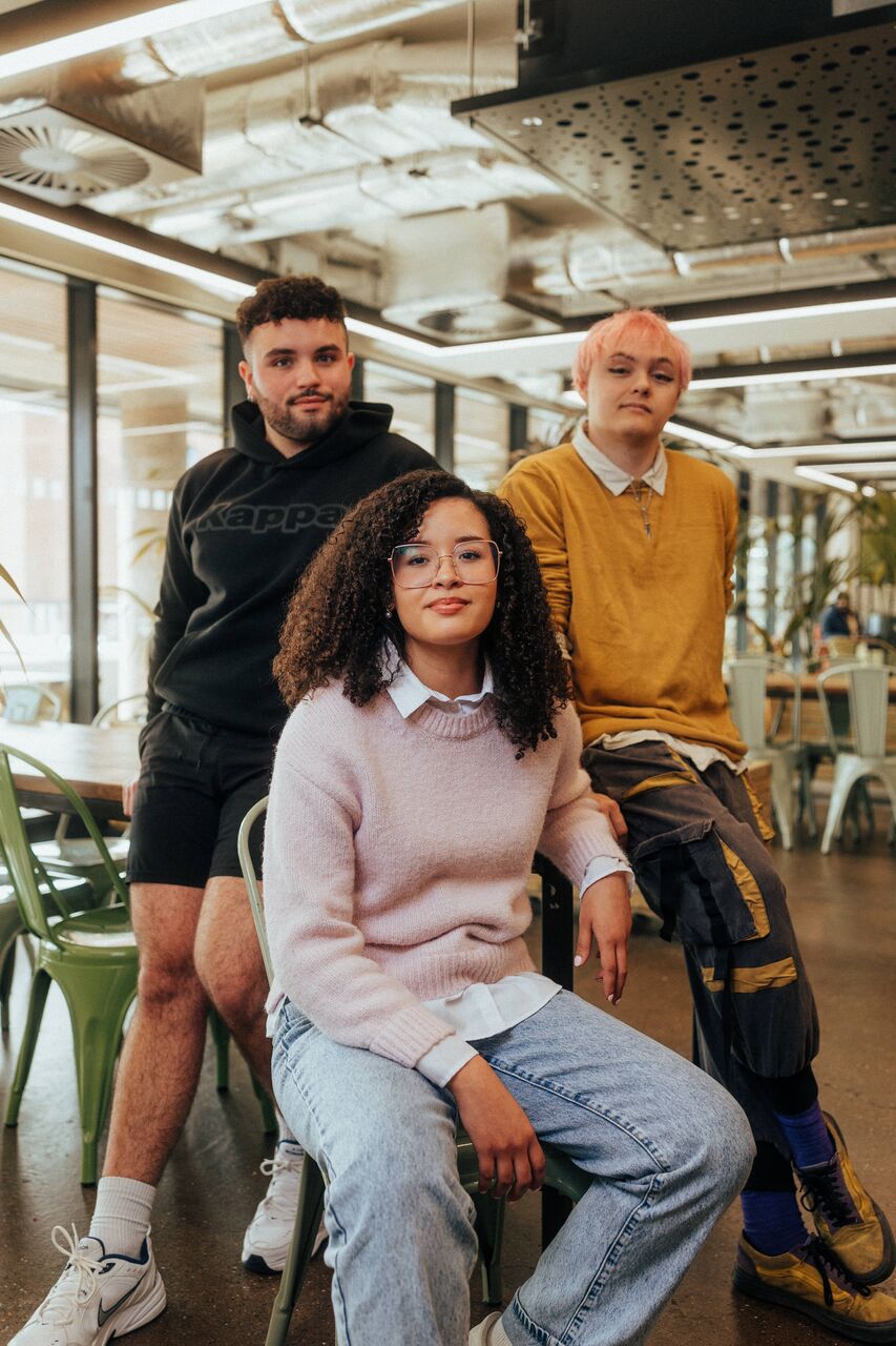 Three of Hull’s students sit confidently together in front of modern lighting and foliage in the student hangout