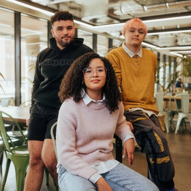 Three of Hull’s students sit confidently together in front of modern lighting and foliage in the student hangout