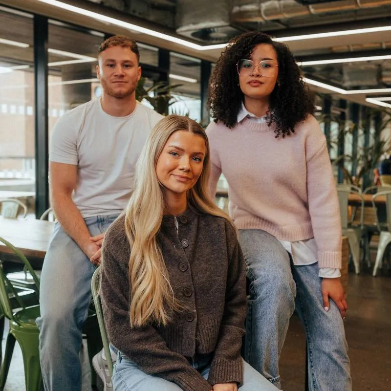 Three of Hull’s students sit confidently together in front of modern lighting and foliage in the student hangout