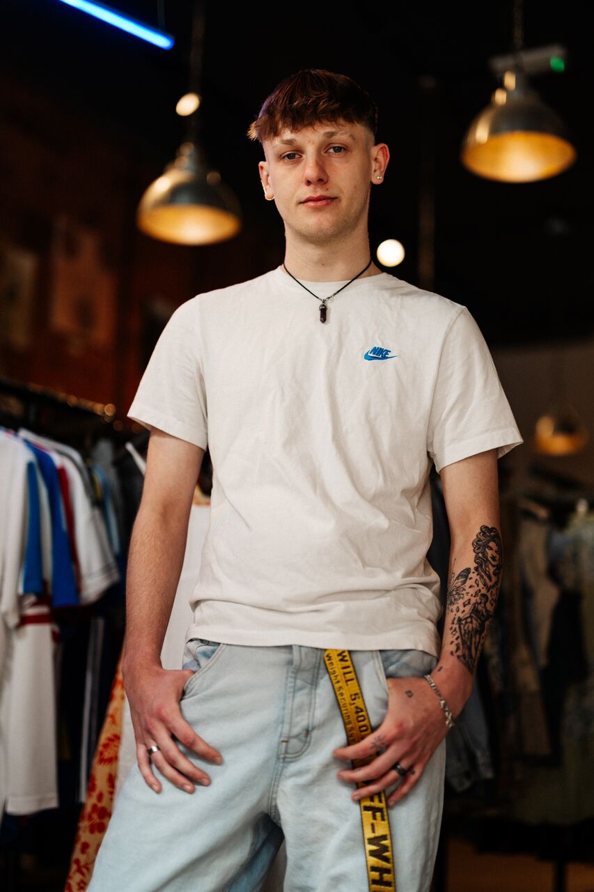A male student in white top and jeans stands confidently under multicoloured strip lighting in a vintage clothes shop