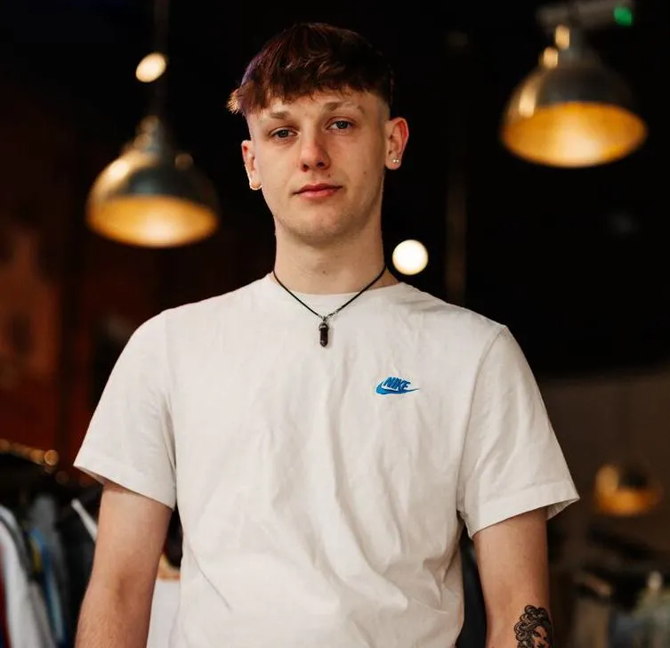 A male student in white top and jeans stands confidently under multicoloured strip lighting in a vintage clothes shop